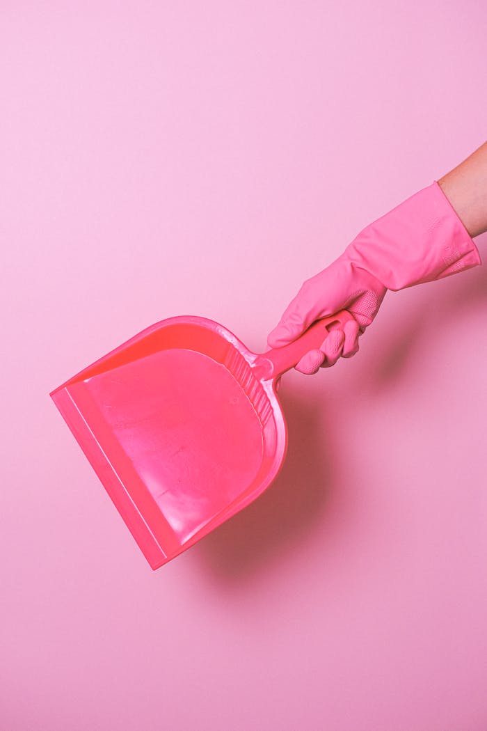 Crop unrecognizable maid in rubber glove showing plastic dust shovel against light pink background in studio